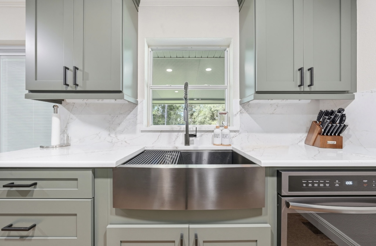Stainless farmhouse apron sink under a window with quartz counters and sage cabinetry
