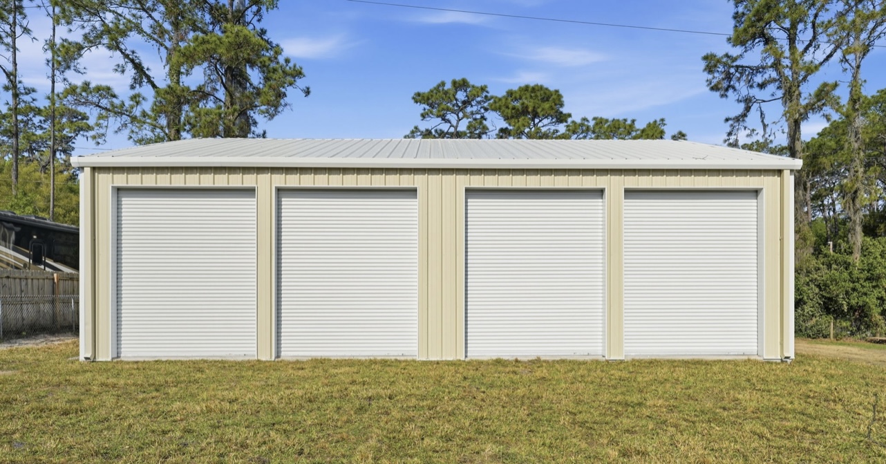 Side elevation of a tan four-bay metal garage with white roll-up doors under Florida pines