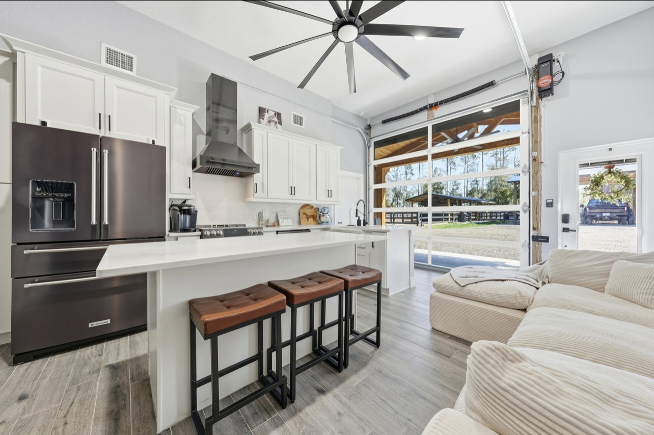 Custom kitchen with white cabinetry, dark appliances and a glass roll-up garage door opening to the property