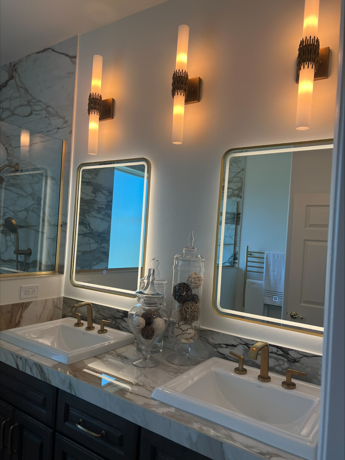 Double vanity with marble counter and backsplash, backlit brass mirrors and matching sconces